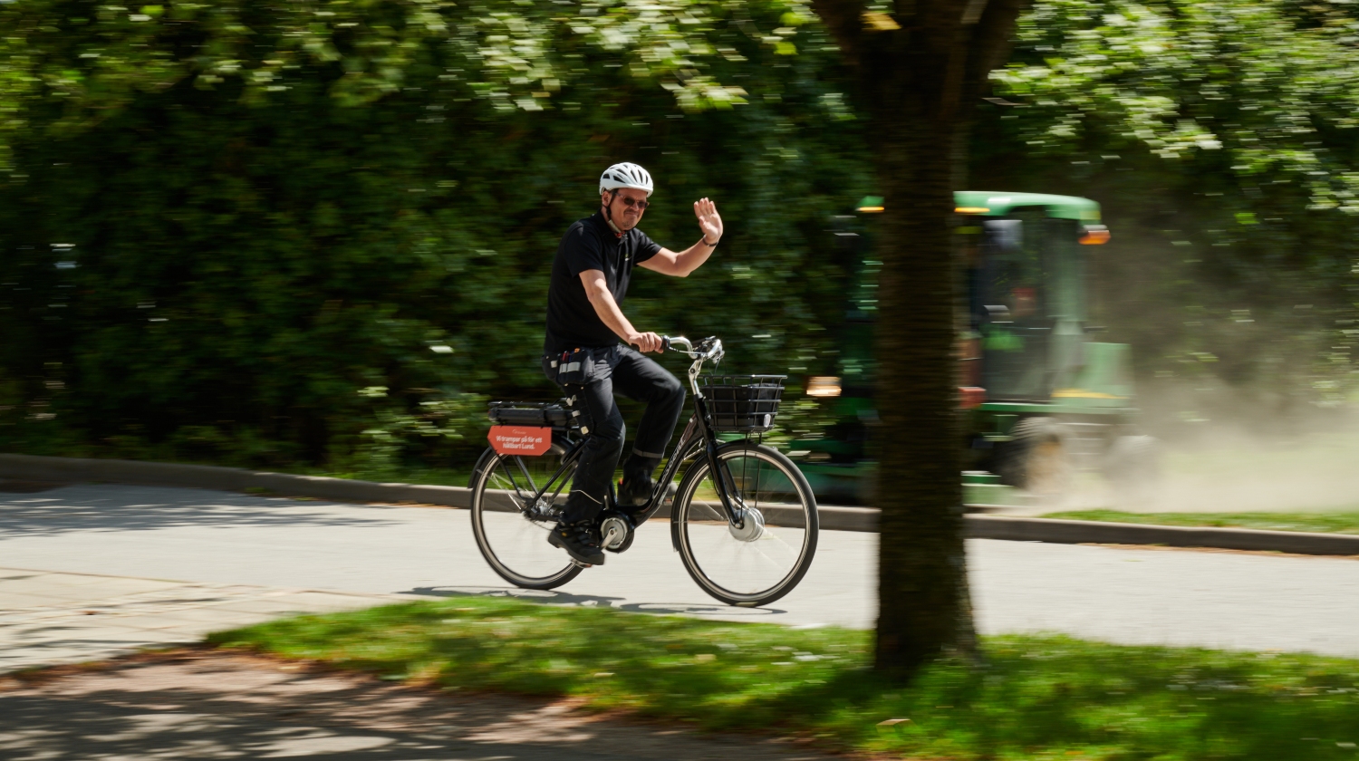Medarbetare som cyklar på en väg kantad av grönska och vinkar till kameran
