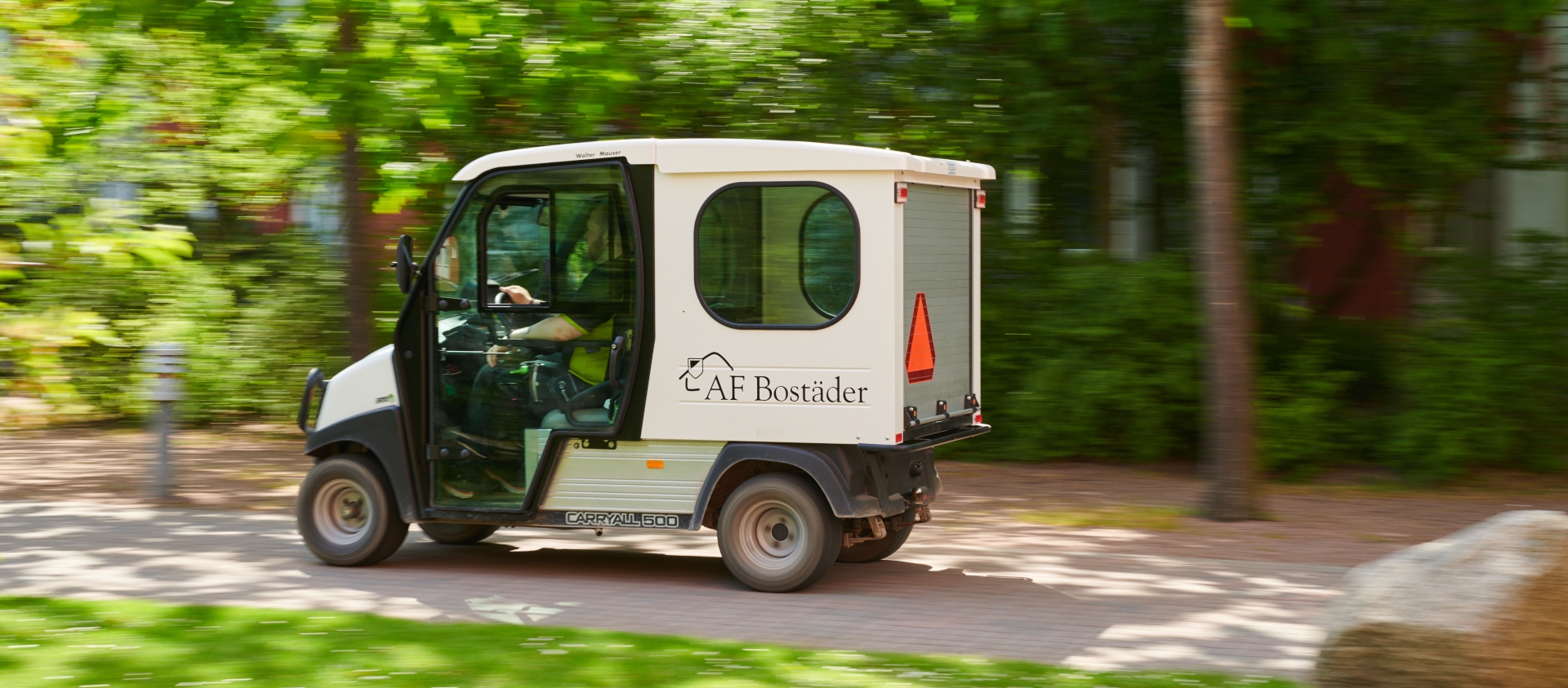 One of AF Bostäder's small electric cars are driving by on a small road surrounded by trees and grass.